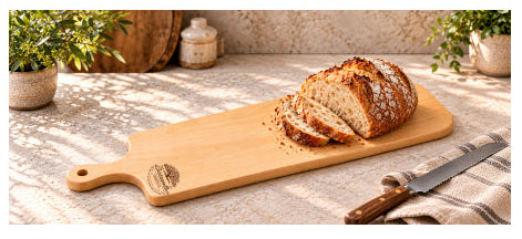 A rectangular wooden cutting board with a handle, shown with a sliced loaf of rustic bread and a knife on a textured stone surface. The scene is styled with potted plants and a wooden barrel in the background.